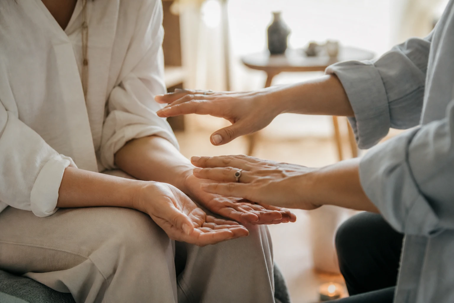 Realistische Nahaufnahme zum Thema energetische Heilung: Hände einer Therapeutin in sanfter, achtsamer Haltung über den Händen einer sitzenden Person, Fokus auf Ruhe, Vertrauen und Berührung ohne direkten Kontakt, weiches natürliches Fensterlicht, warme aber ausgewogene Farbtemperatur, dezente Hintergrundunschärfe, echte Hautstruktur, professionelle Lifestyle-Fotografie, keine übernatürlichen Effekte, kein Kitsch, keine Symbole im Vordergrund und nur sehr subtil im Hintergrund, dezente Atmosphäre, hochwertige Reportagefotografie, natürliche Farben, warme ausgewogene Farbtemperatur, keine magischen Lichteffekte, kein Esoterik-Kitsch, echte menschliche Emotionen und Gelassenheit, realistischer Editorial-Stil